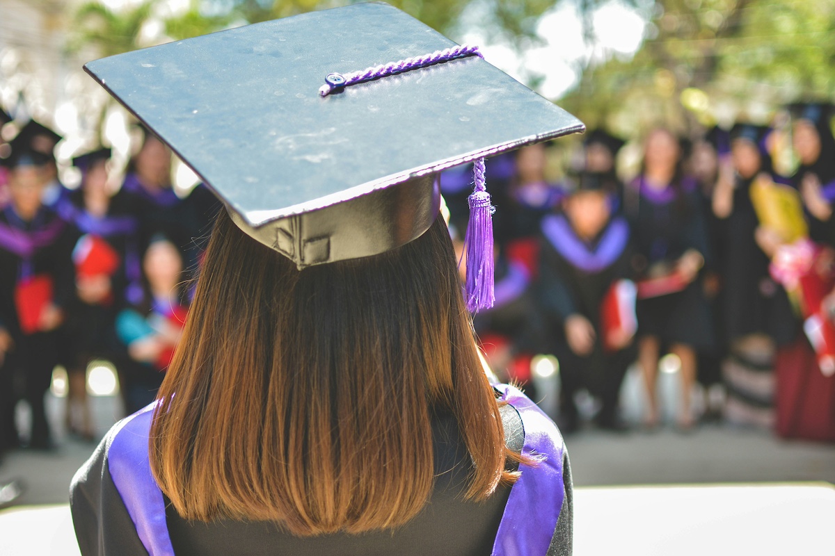 A faceless photo of a female college graduate wearing their graduation cap and robe while looking at their fellow peers