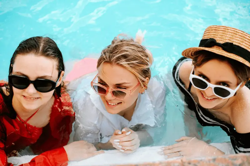 Three women, adorned in sunglasses, lounge on the edge of an aqua pool.