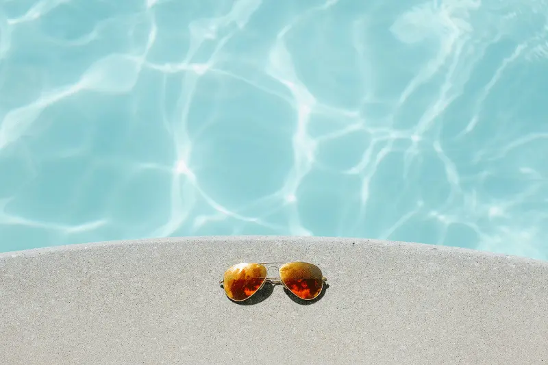 A pair of aviator-style sunglasses lay on a pool deck next to sparkling water.